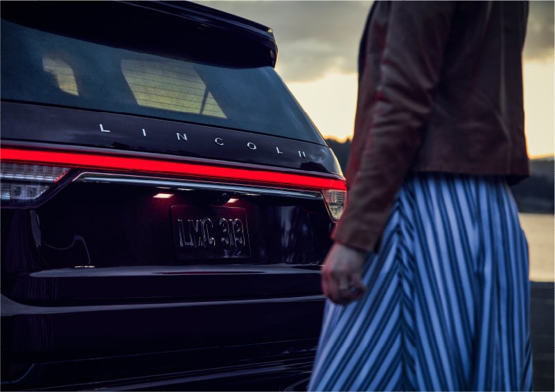A person is shown near the rear of a 2023 Lincoln Aviator® SUV as the Lincoln Embrace illuminates the rear lights | Cogswell Lincoln in Russellville AR