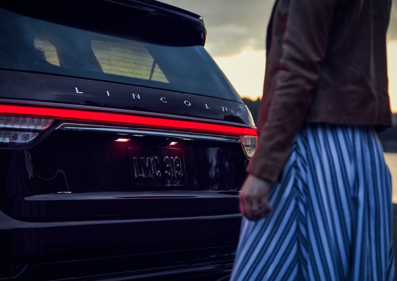 A person is shown near the rear of a 2024 Lincoln Aviator® SUV as the Lincoln Embrace illuminates the rear lights | Cogswell Lincoln in Russellville AR